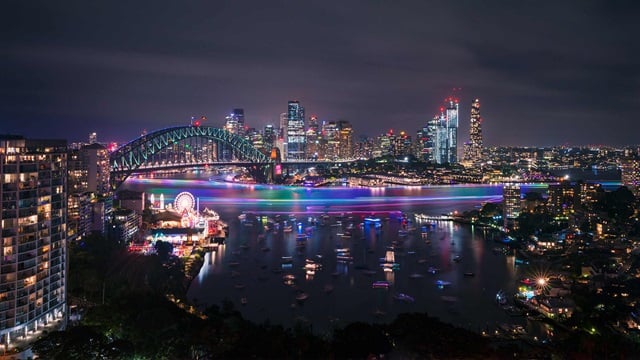 Night view of Sydney Harbour with the illuminated Sydney Harbour Bridge, colorful light trails on the water, and the city skyline in the background.