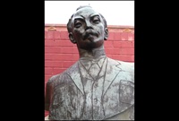 A bronze bust of Jose Marti, mounted on a brick base with a sandstone top against a red painted brick wall.
