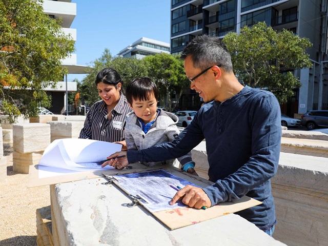 3 people looking at a design on a table, buildings and trees behind them.