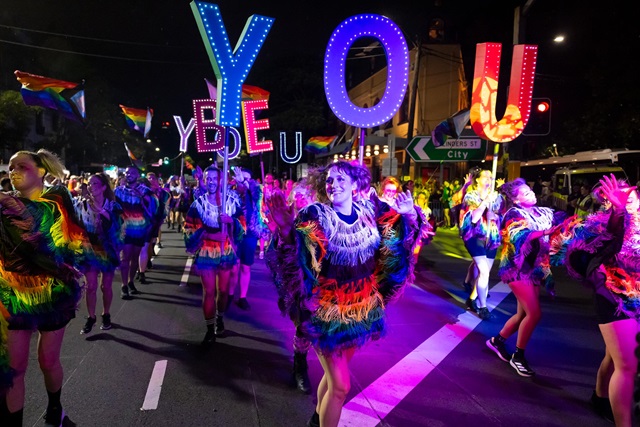 City of Sydney employees wearing rainbow outfits dance together in the evening at a Mardi Gras parade. Some of them hold up letters reading "You be you".