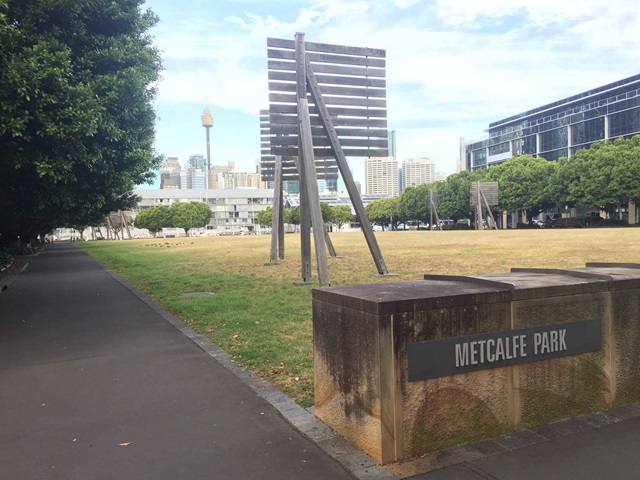 Open grassed area with a view of the city skyline and signage with name of park in the foreground.