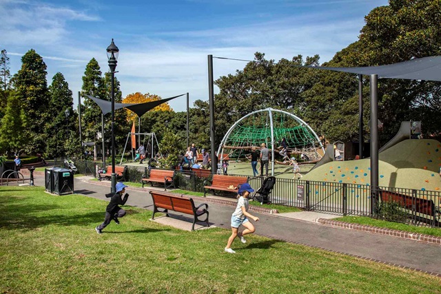 2 children running on grass next to a fenced off playground in a park