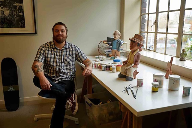 A man with tattoos sits by a desk with art supplies, paint jars, and two sculpted figures, in a well-lit room with a skateboard and framed artwork on the wall.