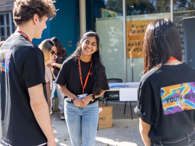A group of individuals standing outside a building on a sunny day. One person faces the camera, wearing a black T-shirt and blue jeans, with a lanyard. Two others are seen from the side, engaged in conversation. They wear T-shirts with “Youth Xpress” written on them. Behind them, there’s a table with papers and boxes.