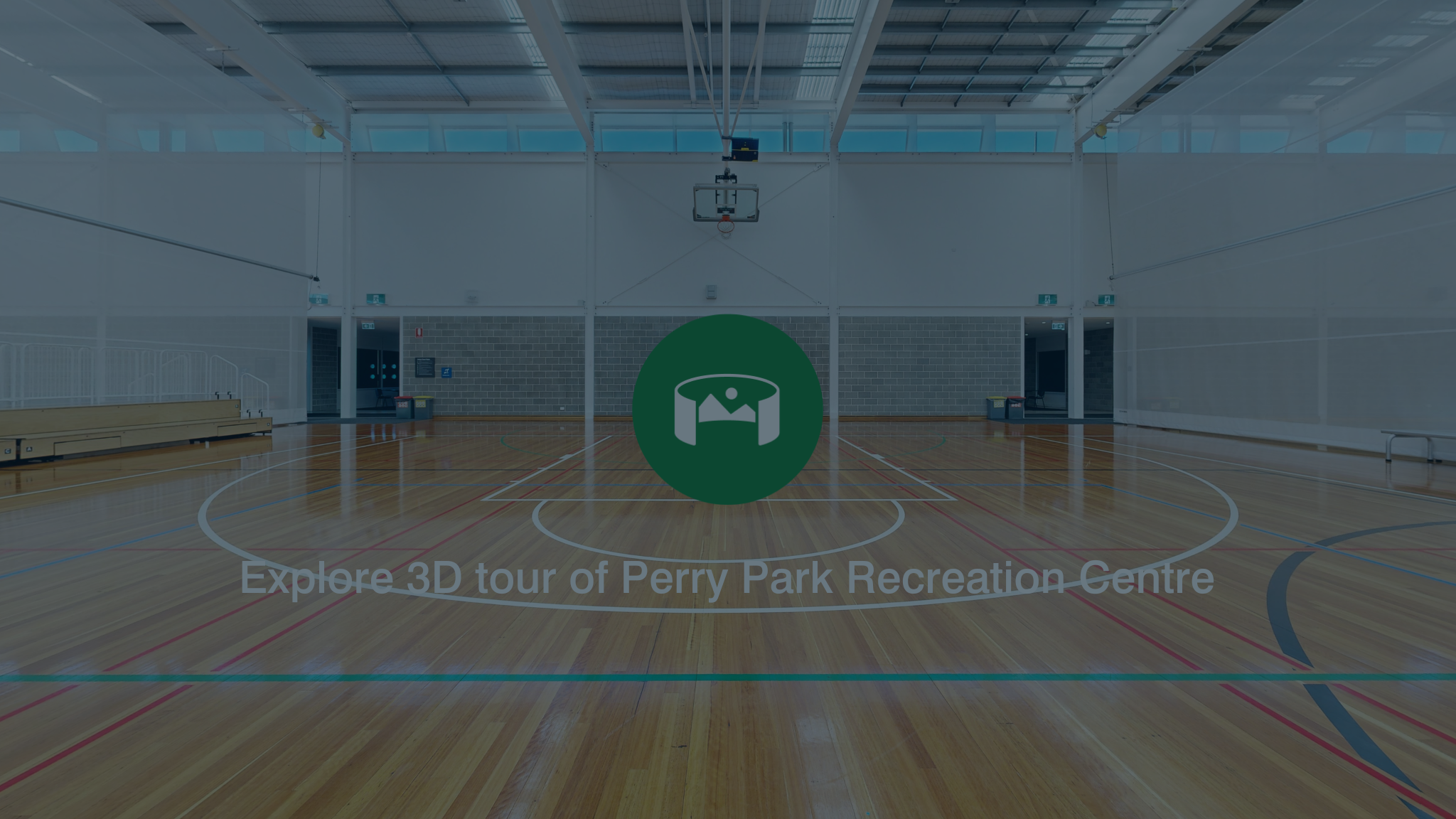 Interior view of an empty indoor basketball court with wooden flooring, surrounded by high walls and a ceiling with windows with the text explore 3D tour of Perry Park Recreation Centre