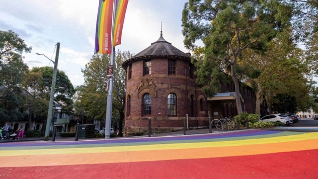 Rainbow painted on a city street with a round brick building and rainbow flag in the background